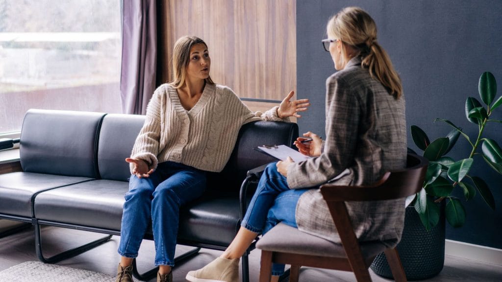 Two women sitting across from each other in an office, one holding a clipboard, engaged in conversation.