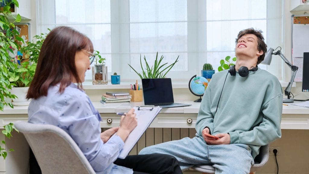 A person with headphones around their neck leans back in a chair, smiling, while another person opposite writes on a clipboard. A desk with plants and a laptop is in the background.