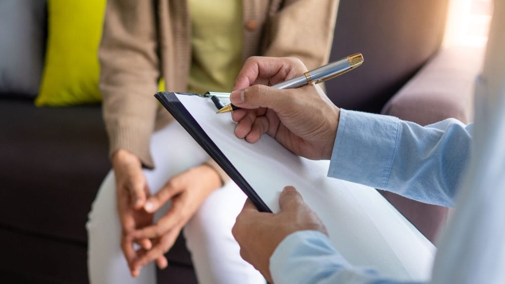 A therapist writes notes on a clipboard while a patient sits nearby, engaged in a counseling session in a comfortable setting.